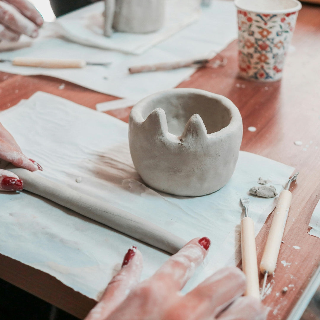Clay pot on a table with pottery tools and a floral-patterned cup in the background