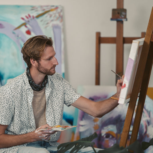 Man painting on an easel in a studio with colorful artworks in the background